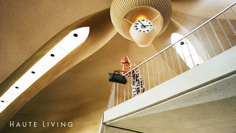 Woman in dress standing on a landing with metal railing holding a handbag below a ceiling with lights and a large clock fixture.