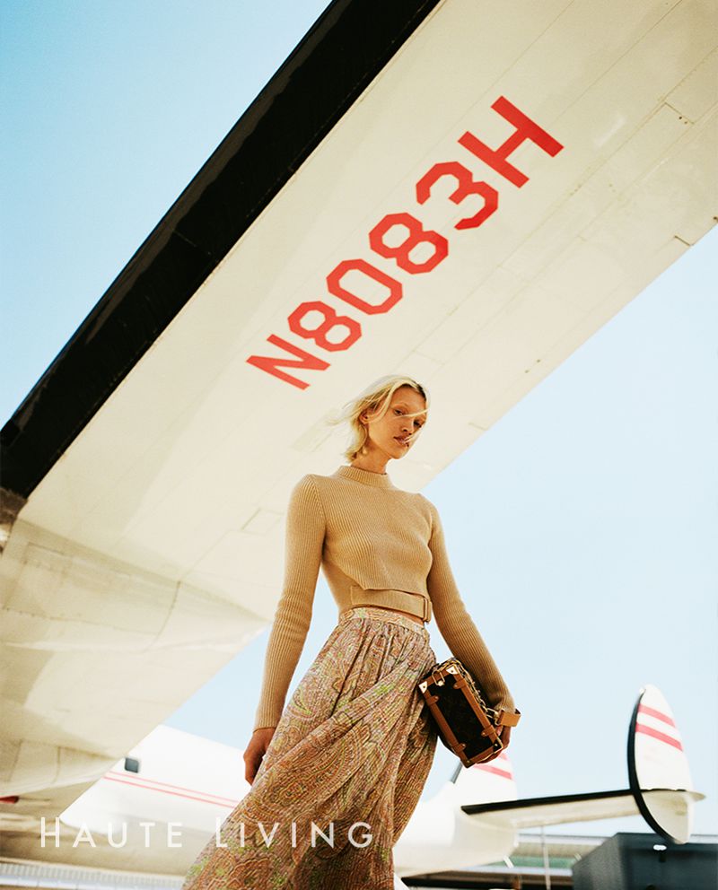 A woman in a dress stands in front of an airplane, holding a handbag. Airplane wing has red letters, a blue sky is behind the plane.