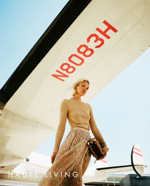 A woman in a dress stands in front of an airplane, holding a handbag. Airplane wing has red letters, a blue sky is behind the plane.