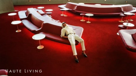 Model wearing a white shirt and trousers reclining on a curved red lounge bench in a modern interior space