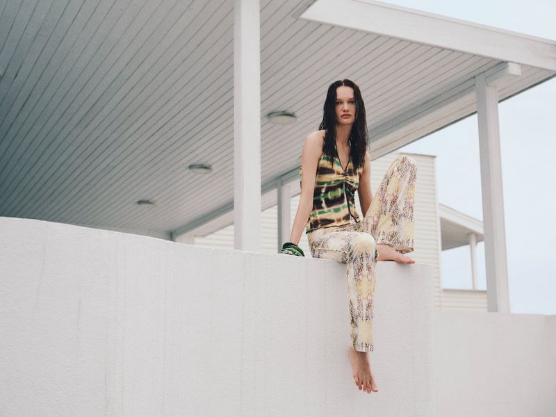 Brunette model in a tie-dye top and patterned pants sits barefoot on a white ledge in front of a white building on an overcast day.