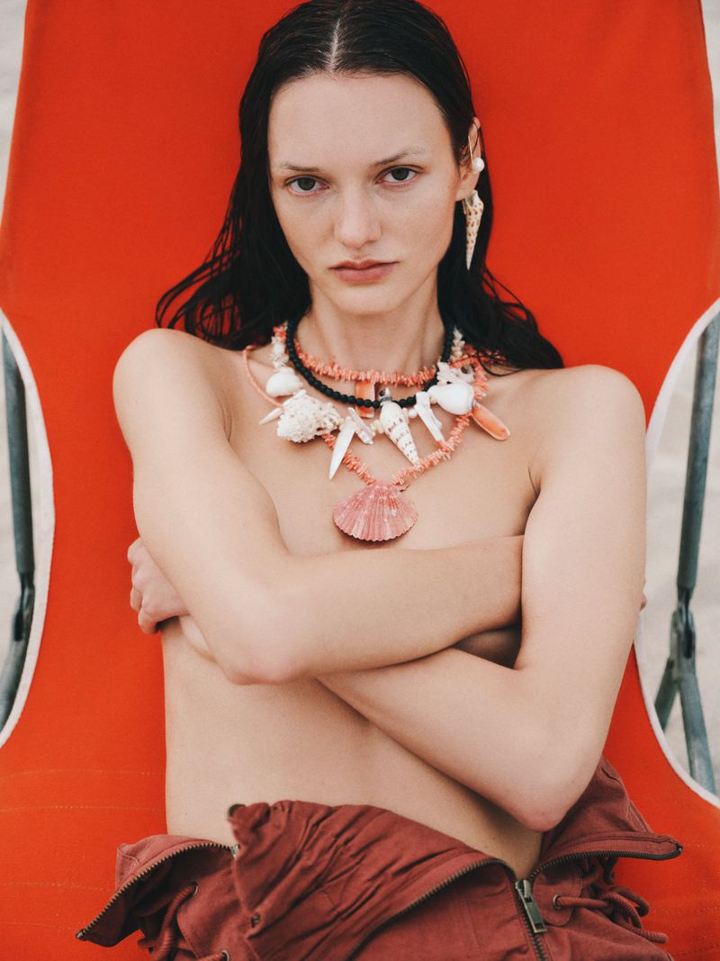 A model poses on a beach chair wearing a shell necklace and earring, with a close-up shot emphasizing summer fashion and seaside aesthetics.