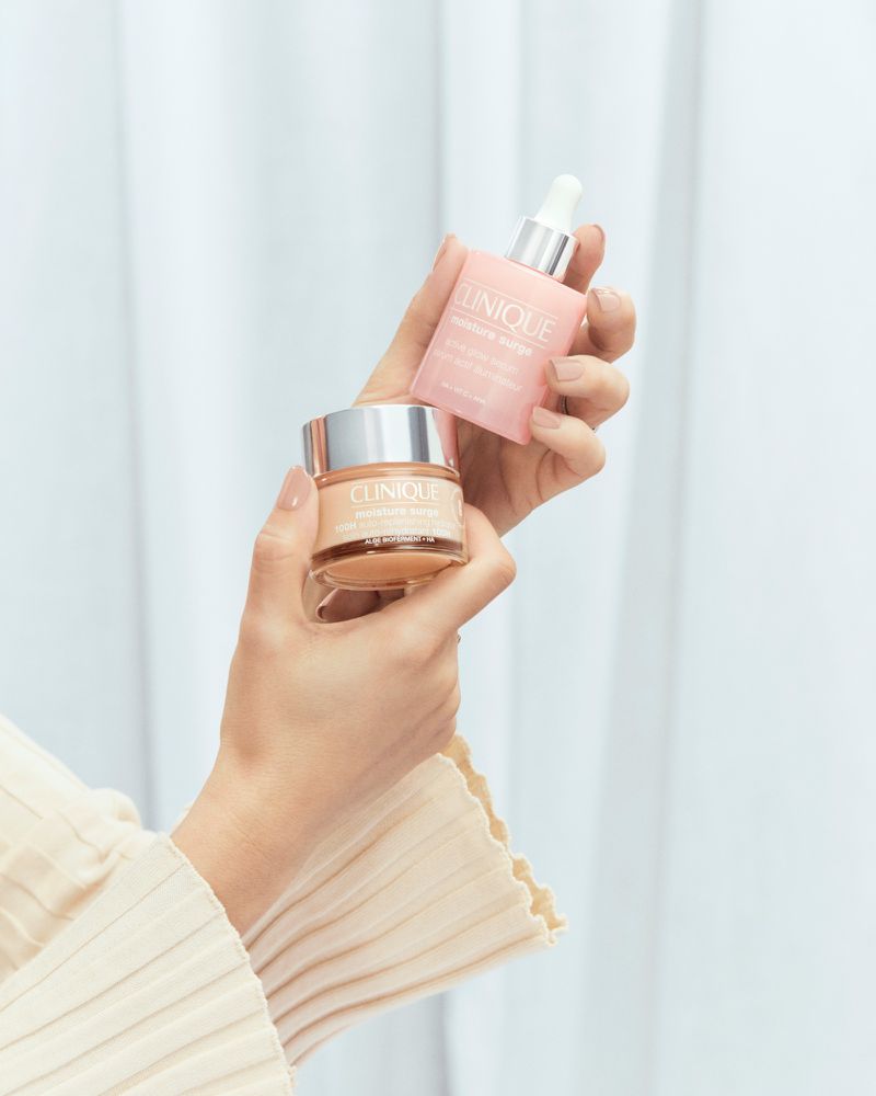 Two hands holding a Clinique Moisture Surge moisturizer jar and a hydrating supercharged concentrate serum bottle in an indoor setting.