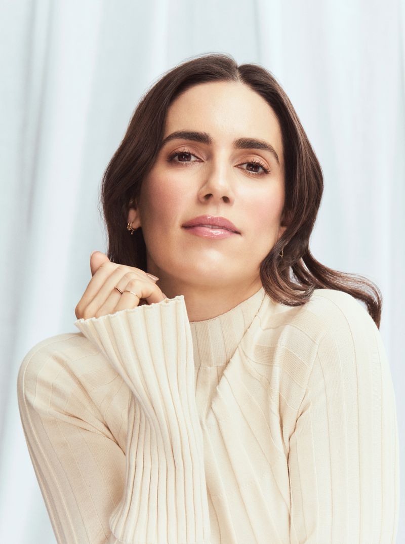 Portrait of a brunette woman wearing a cream-colored ribbed sweater, looking directly at the camera against a soft, light blue background.