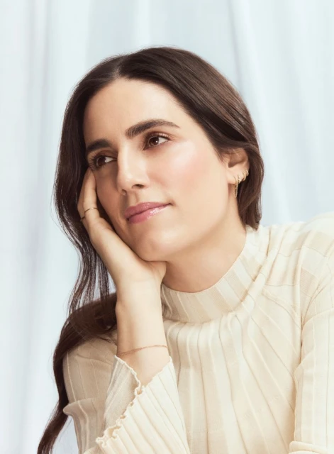 Close-up shot of a brunette woman in a white top, resting her head on her hand, in a naturally lit indoor setting with a serene expression.