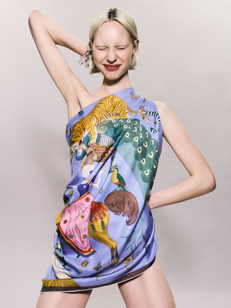 Woman poses in printed silk scarf dress with tiger, peacock, and butterfly pattern in studio lighting on neutral background.