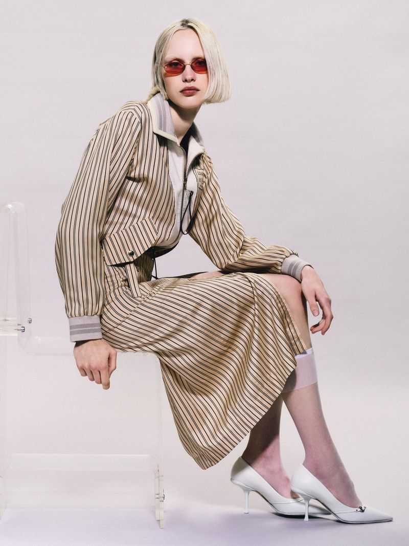 A fair-skinned young adult female in a striped suit poses on a transparent chair in a studio, wearing red sunglasses and white heels.