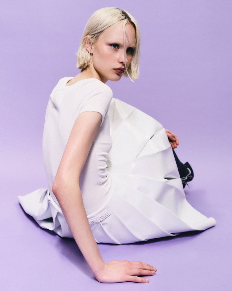 Woman with platinum blonde hair sits in studio wearing white outfit against lavender background. Fashion and beauty shot with neutral expression.