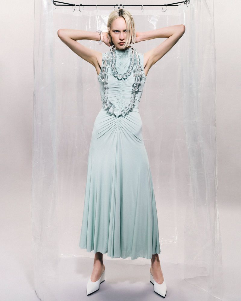 A full shot of a female fashion model in a light blue dress and white wedge heels stands in front of a translucent curtain.