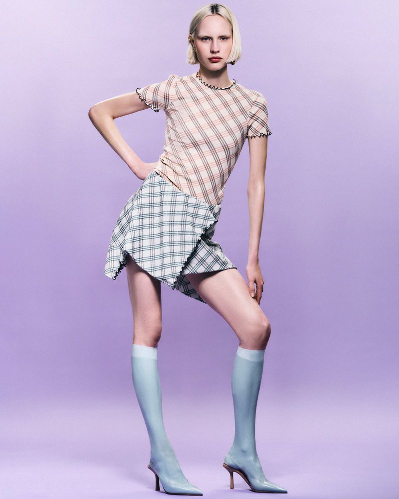 Fashion model poses in a studio wearing a checkered top and skirt, knee high socks, and high heels, with a solid lavender background.