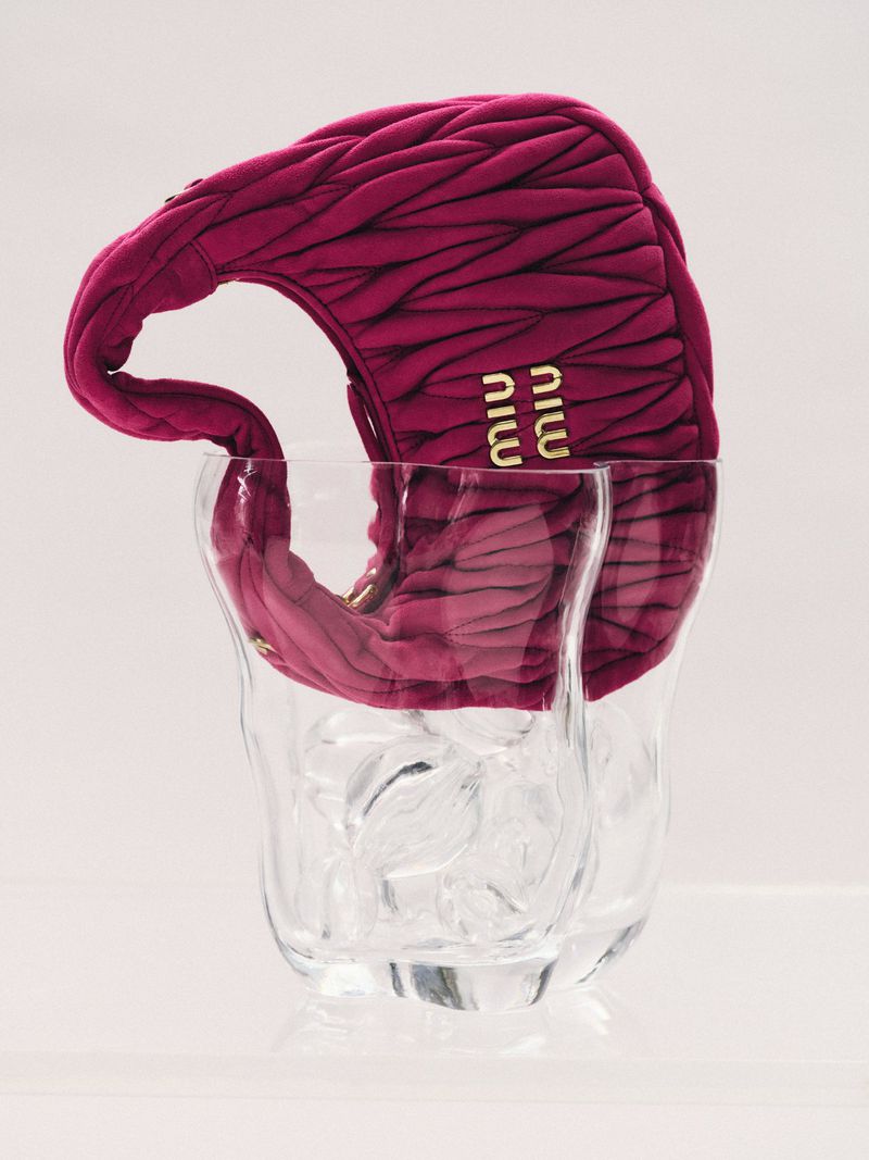 Burgundy Miu Miu purse in a clear glass vase against a white background. Elegant fashion accessory with a luxury aesthetic.