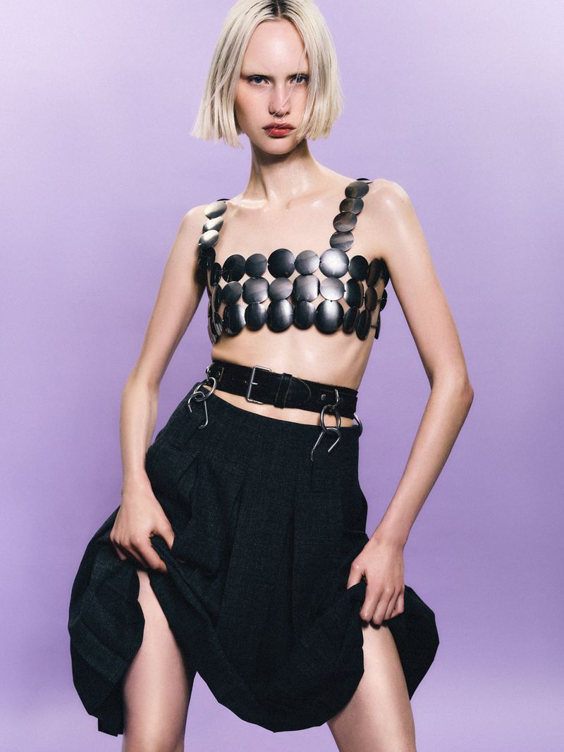 Fashion model in a metal bra top and black skirt strikes a pose against a lilac backdrop in a studio shot, showcasing trendy apparel.