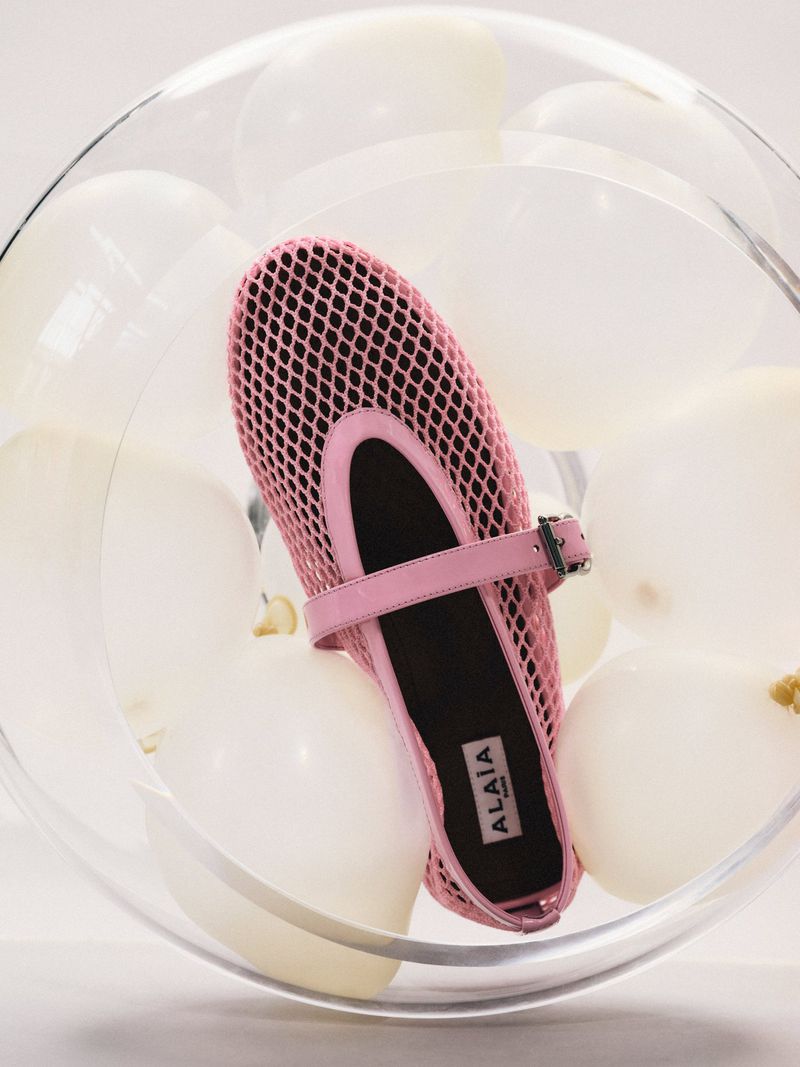 Close-up studio shot of a pink Alaïa ballet flat inside a clear glass bowl filled with white balloons against a white backdrop.
