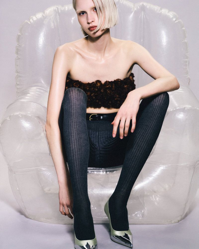Studio shot of a fair-skinned model sitting on an inflatable chair, wearing a brown strapless top, gray leggings, and silver heels.