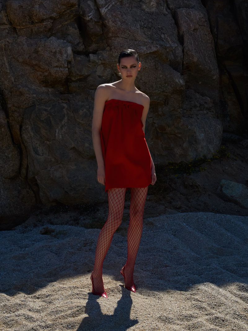 A woman in a red dress and fishnet stockings poses in a desert environment against a rocky backdrop in an outdoor fashion shot.