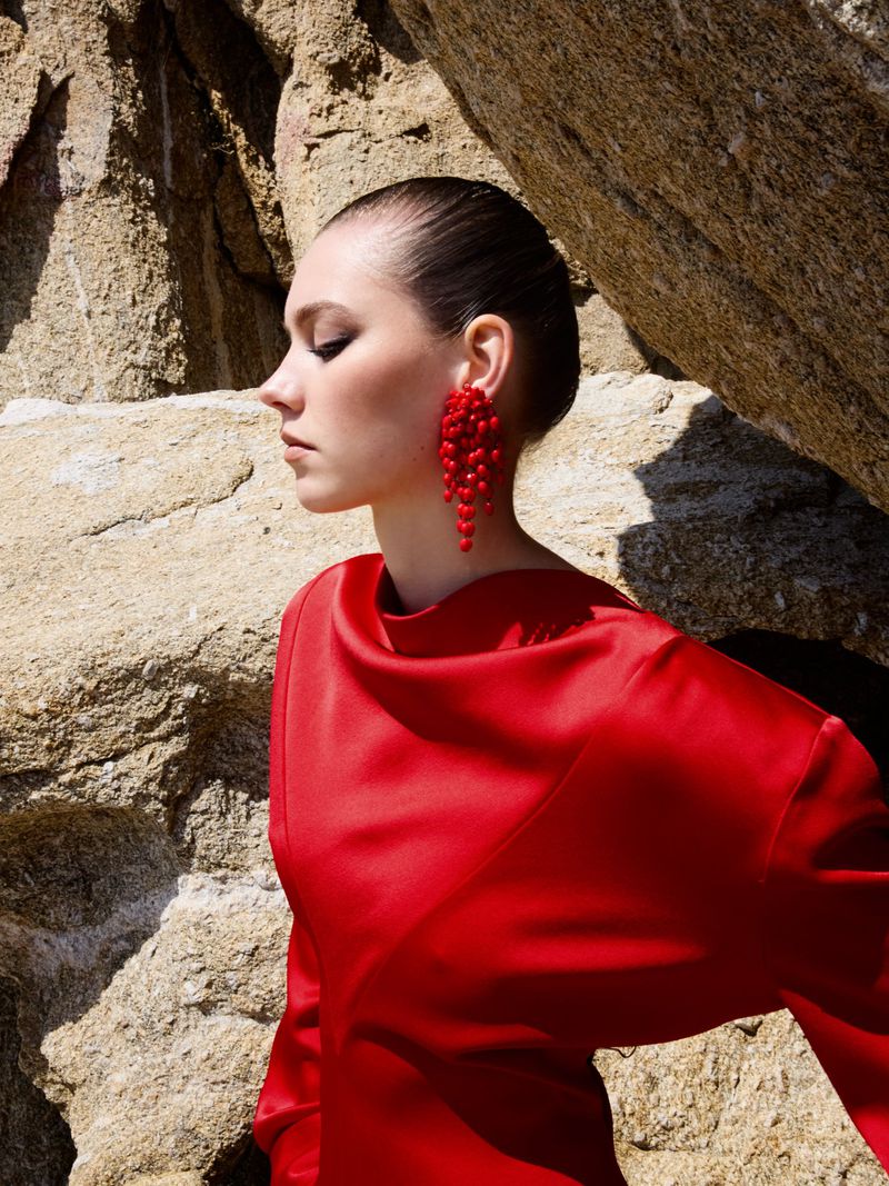 Woman in red dress with red earrings posing against a rock formation. Fashion shot with natural lighting and a side view.