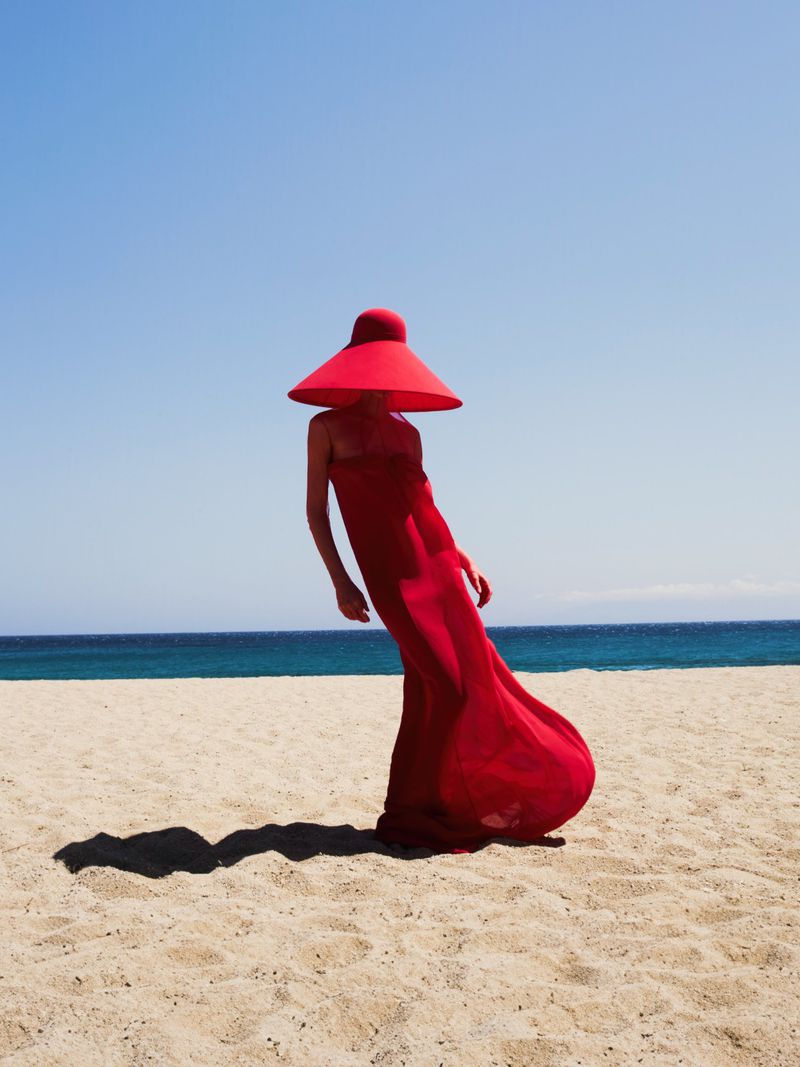 Woman in flowing red gown and wide-brimmed hat stands on a sandy beach with the ocean and blue sky in the background.