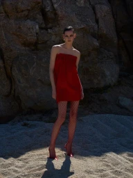 Model standing on sandy ground in a strapless red dress and fishnet tights against a rocky background