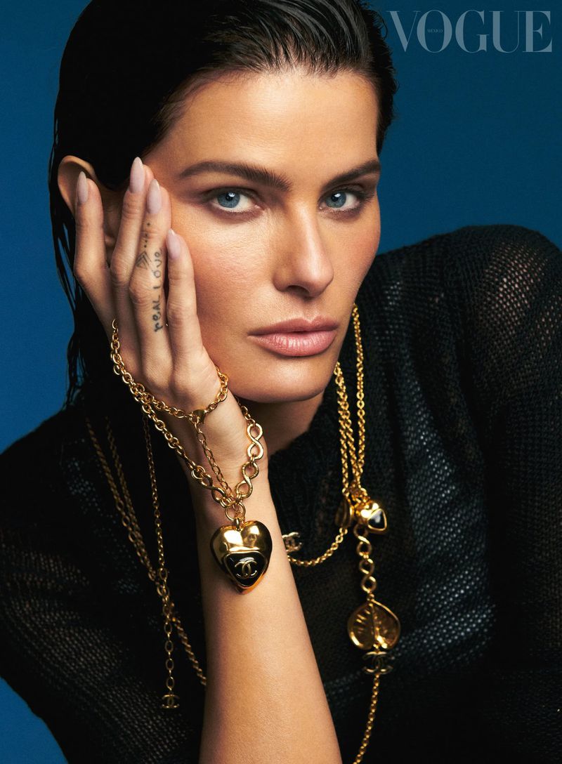 Close-up fashion portrait of a woman wearing gold jewelry, black top, and posing against a blue background for a Vogue magazine cover.