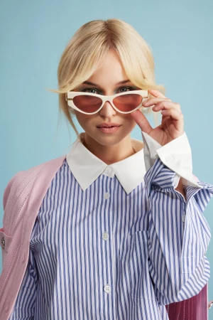 Blonde woman wearing white cat-eye sunglasses and a blue and white striped button-down shirt against a solid blue background.