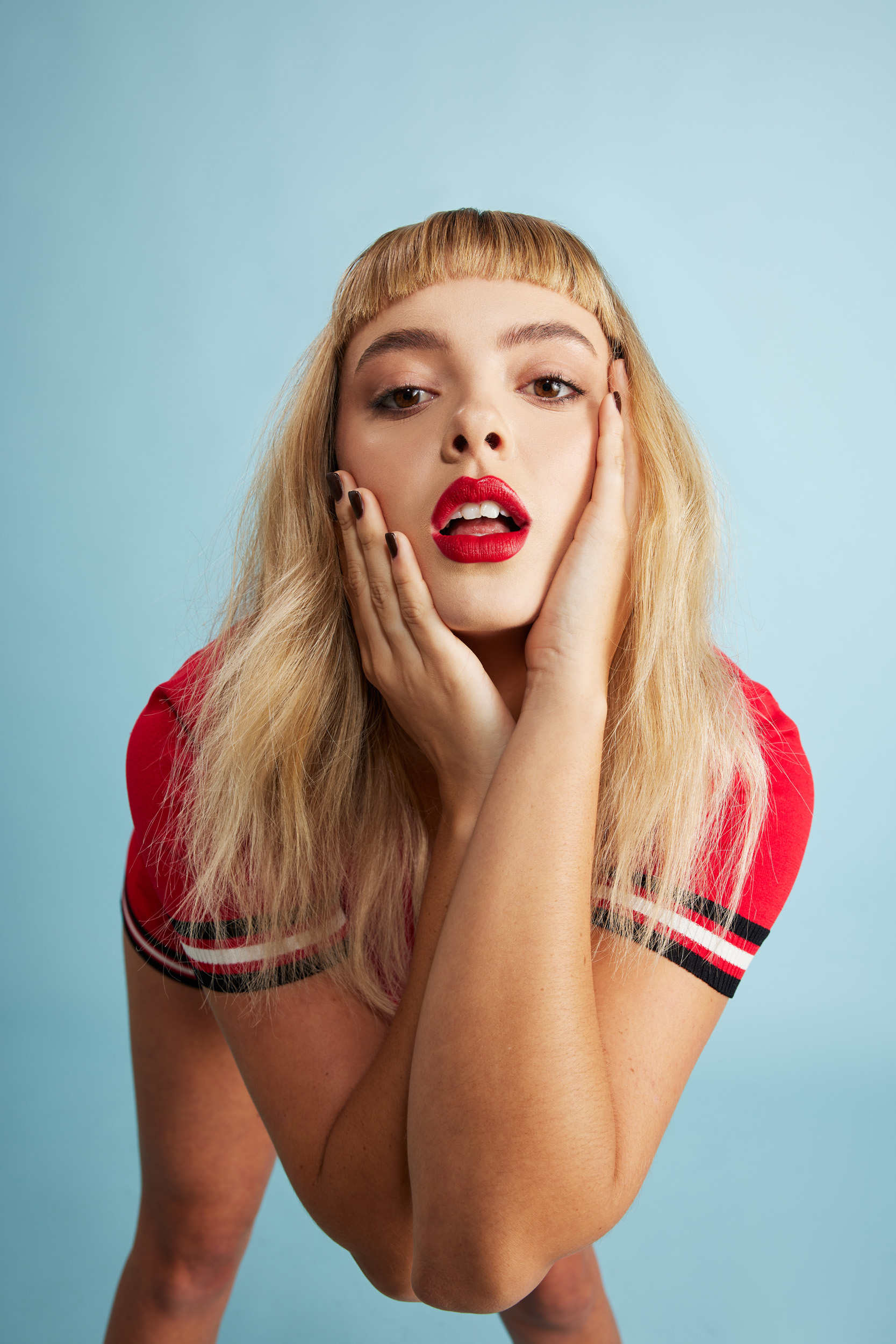 Blonde woman with hands on her face, wearing a red shirt, and makeup posing against a blue backdrop in a beauty shot.