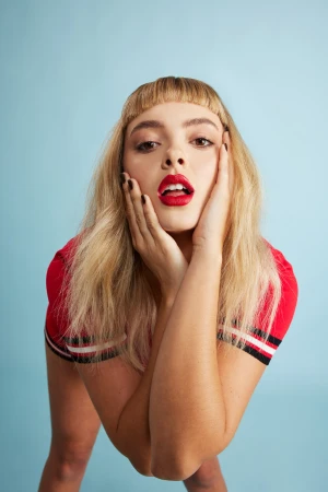 Close-up portrait of a woman with blonde hair, bangs, and bold red lipstick holding her face with both hands against a blue background.