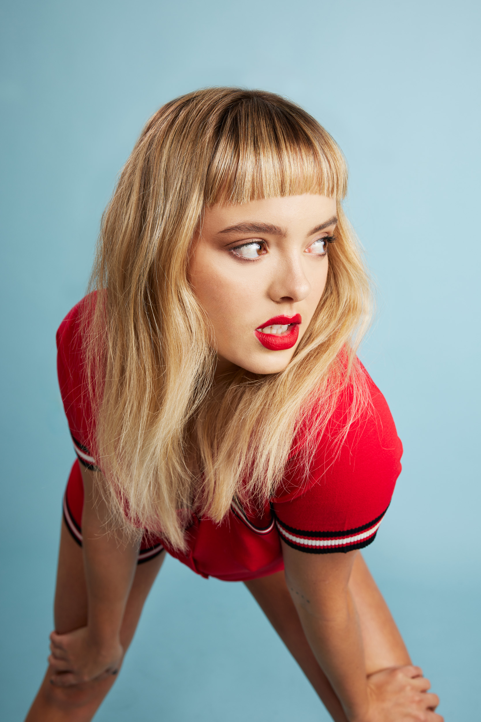 Blonde woman with bangs and red lipstick poses in a red baseball jersey against a light blue background, looking away.