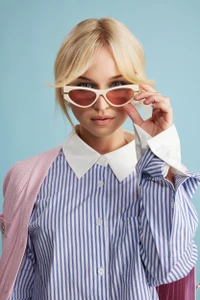 Blonde woman wearing white cat-eye sunglasses and a blue and white striped button-down shirt against a solid blue background.