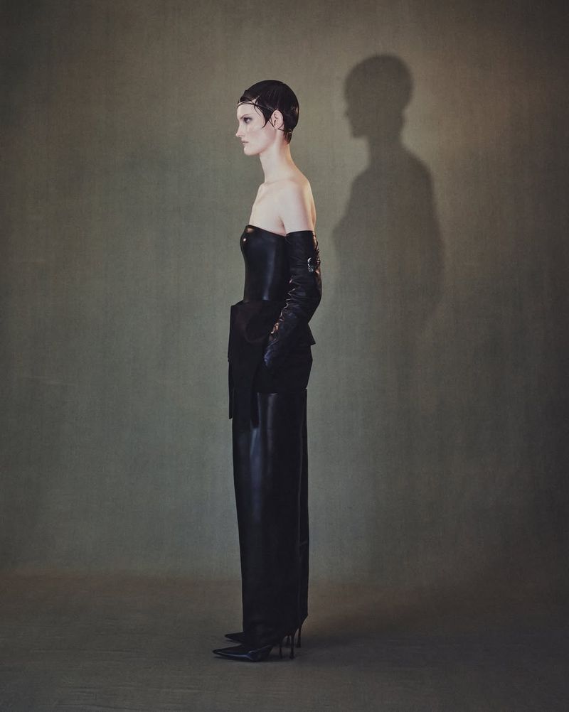 Elegant woman in black strapless dress, long gloves, and heels poses in a fashion shot with an olive green background and visible shadow.