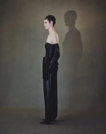 Elegant woman in black strapless dress, long gloves, and heels poses in a fashion shot with an olive green background and visible shadow.