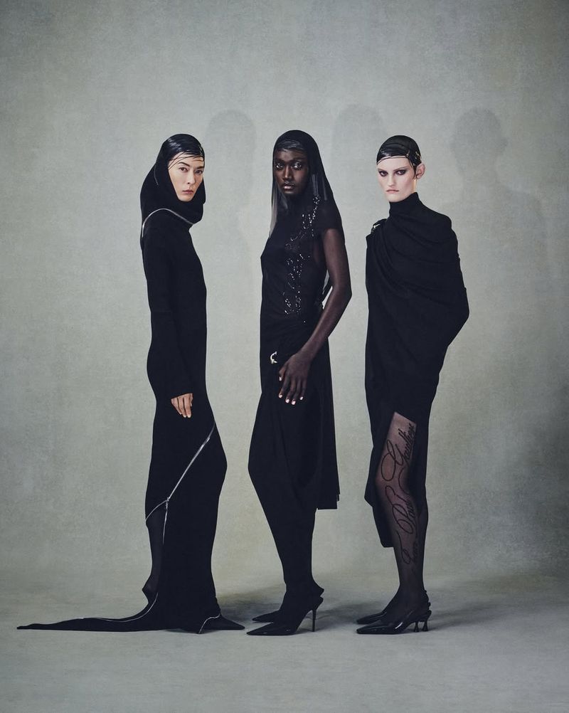 Three women pose in black hooded outfits in a studio setting. The models stand in a line against a gray backdrop in a fashion editorial.