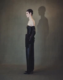 Model in profile wearing a black strapless gown and gloves with a sharp shadow cast on a textured wall.