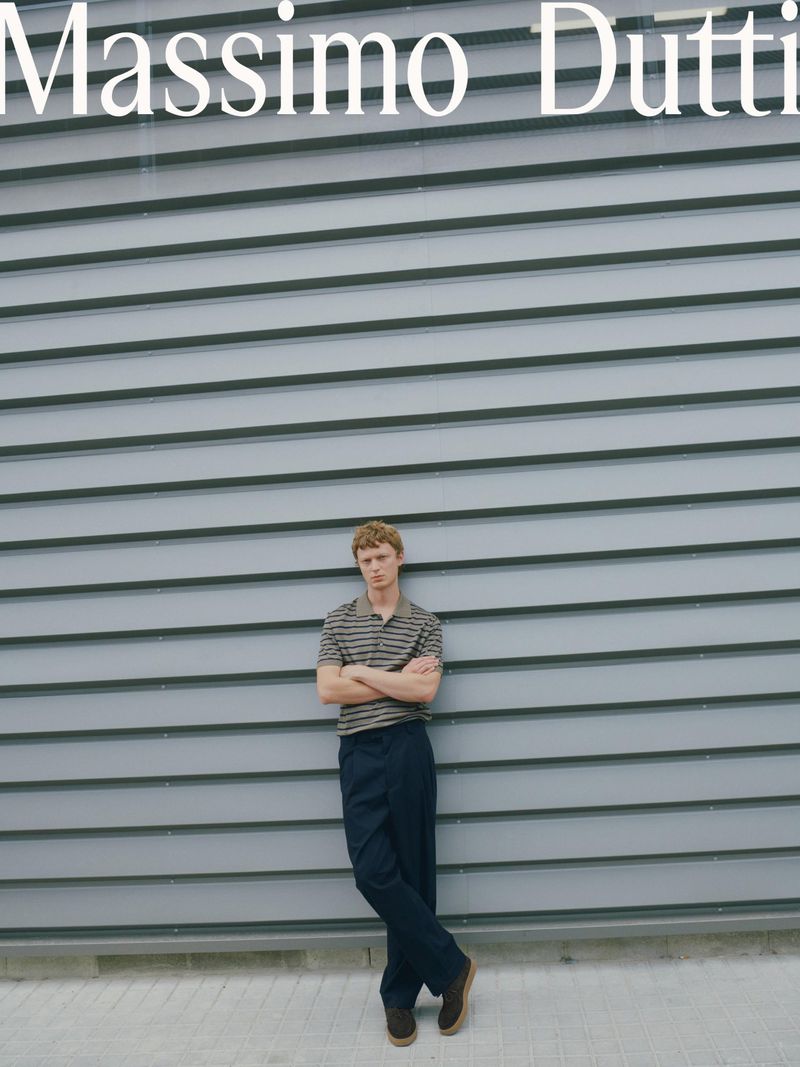A young adult male leans against a gray wall wearing a striped shirt and dark pants, showcasing a relaxed, casual fashion style outdoors.