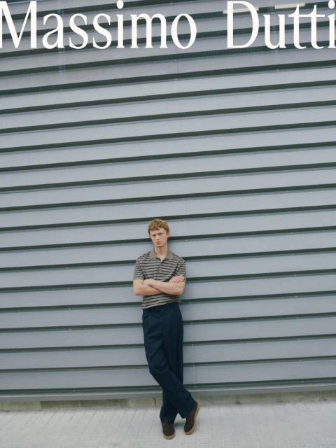 A young adult male leans against a gray wall wearing a striped shirt and dark pants, showcasing a relaxed, casual fashion style outdoors.