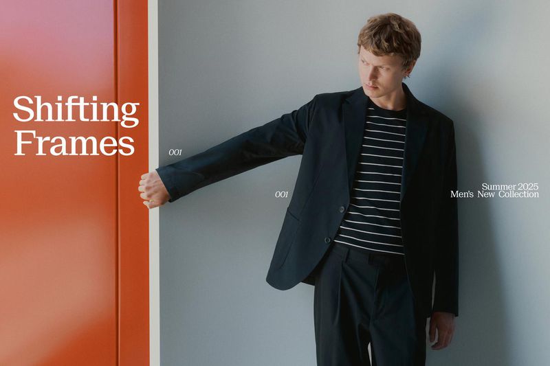 Mens fashion shoot featuring a male model in a black suit and striped shirt leaning against a gray wall with an orange panel.