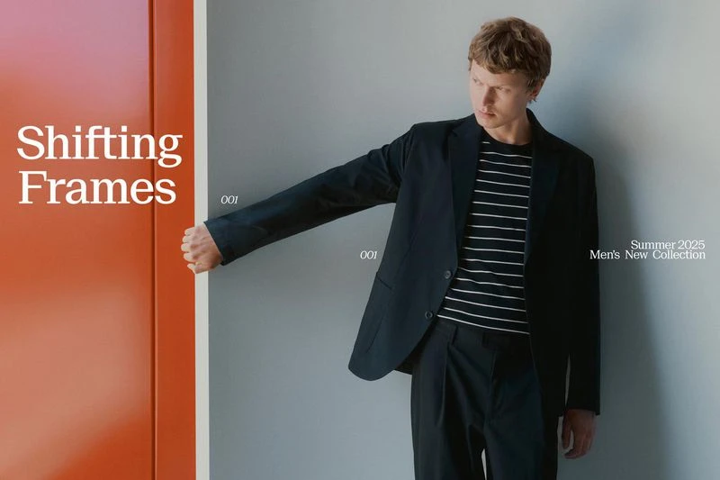 Mens fashion shoot featuring a male model in a black suit and striped shirt leaning against a gray wall with an orange panel.