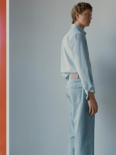 Studio shot of a fair-skinned adult male in a light blue denim shirt and jeans, posing in profile against a gray background.