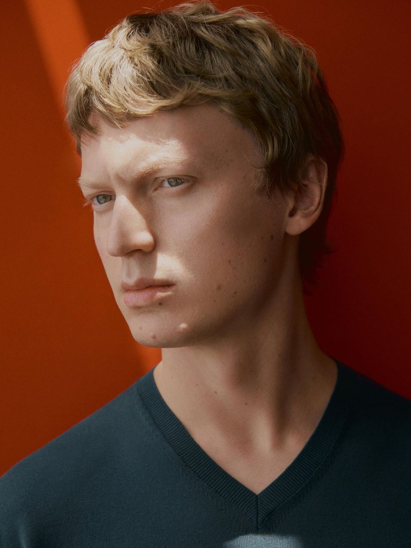 Portrait of a light-skinned young man with blond hair wearing a dark blue sweater against an orange background with soft window light.