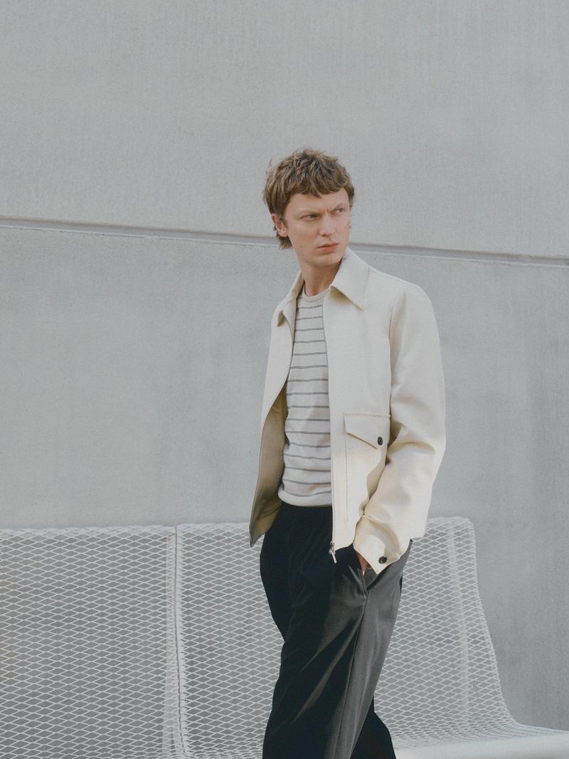 A young adult man in a light jacket and dark pants stands outdoors against a gray wall with a metal bench for a fashion shot.