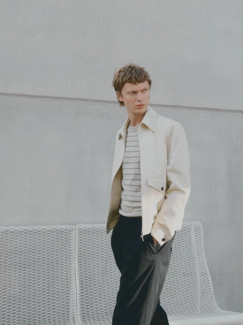 A young adult man in a light jacket and dark pants stands outdoors against a gray wall with a metal bench for a fashion shot.