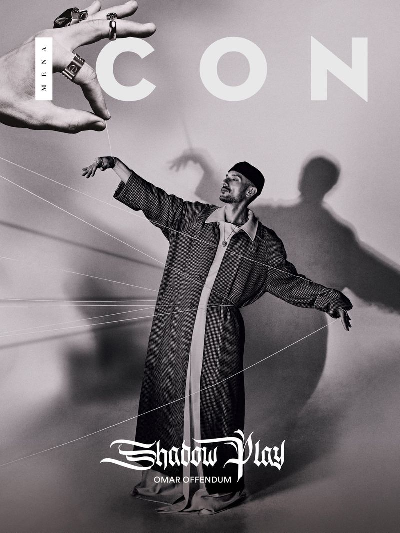 A man in a long coat posed under an oversized hand with strings attached, evoking a puppet show, shot in black and white for ICON magazine.