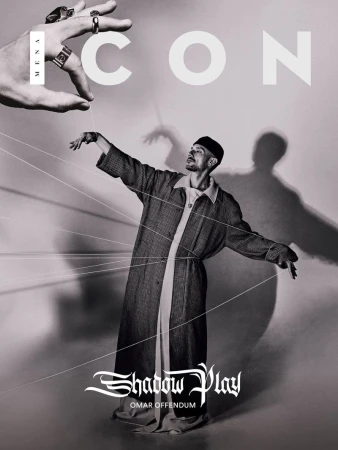A man in a long coat posed under an oversized hand with strings attached, evoking a puppet show, shot in black and white for ICON magazine.