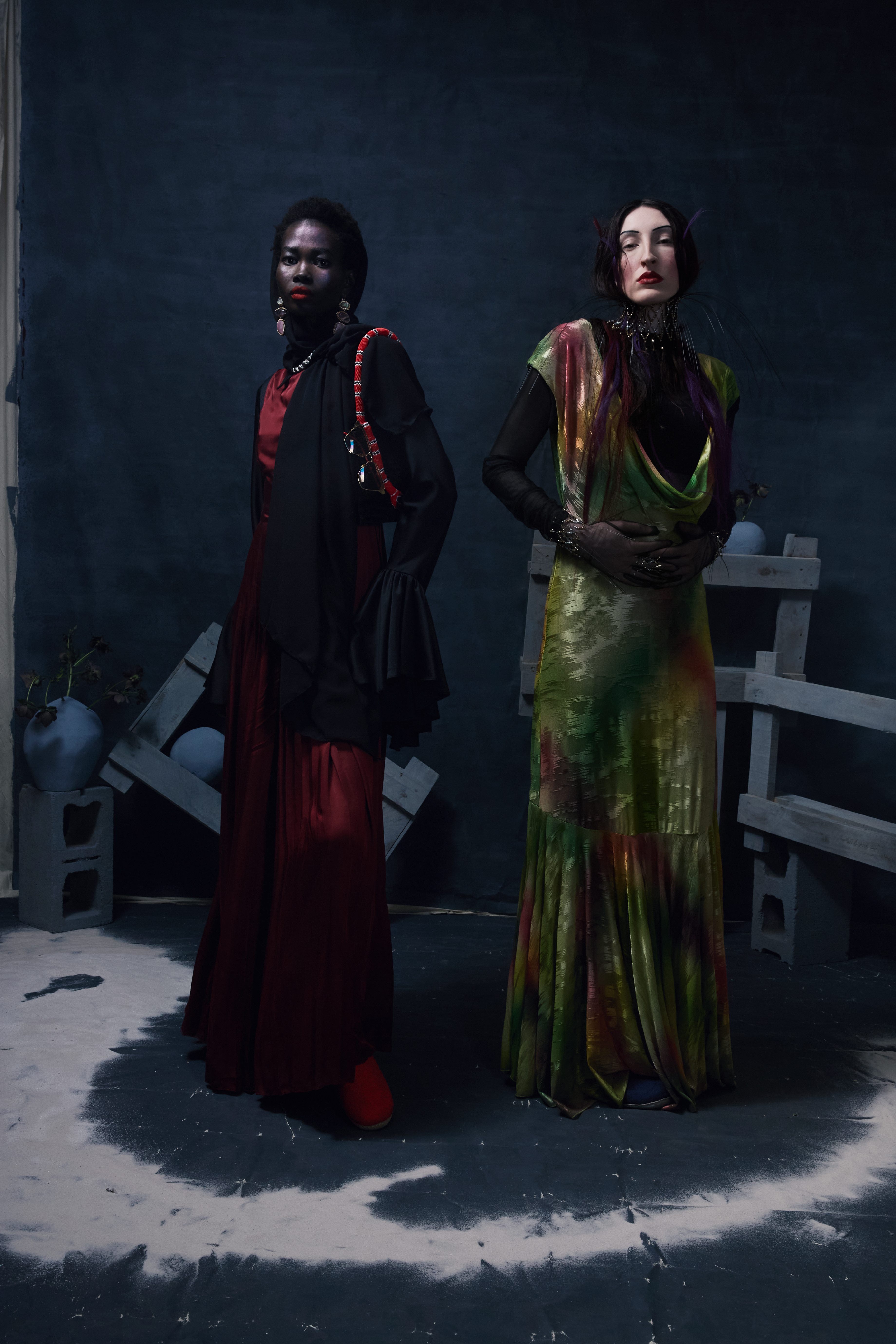 Two women in elegant gowns pose in a studio with a dark backdrop, concrete blocks, and vintage-inspired styling for a fashion editorial.