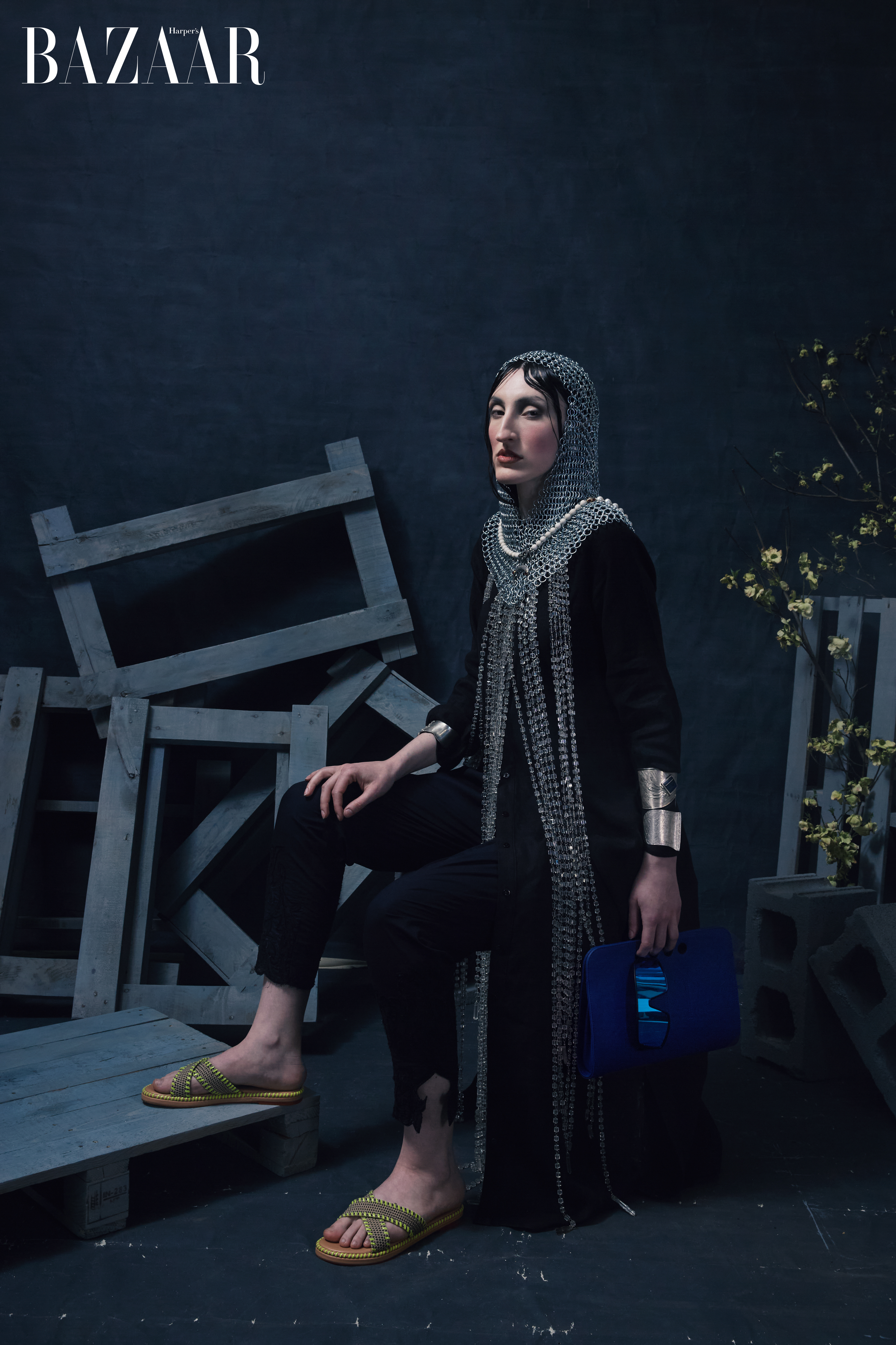 A fashionable adult female sits wearing a chainmail hood and black robe, in a gritty studio environment with wooden pallets and sparse plants.