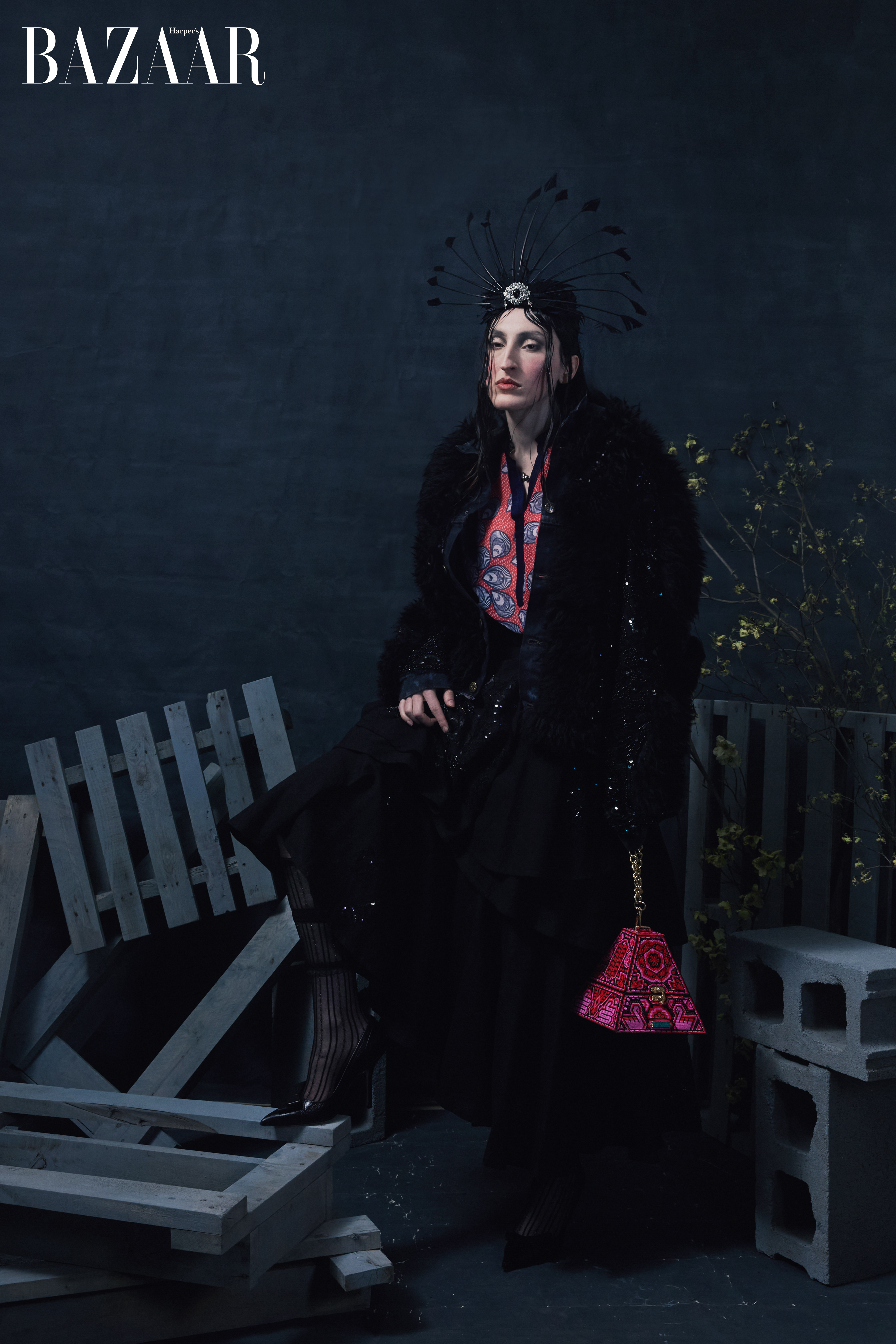 A woman in an extravagant outfit sits against a dark backdrop, featuring a patterned top, ornate headdress, and a sequined coat.