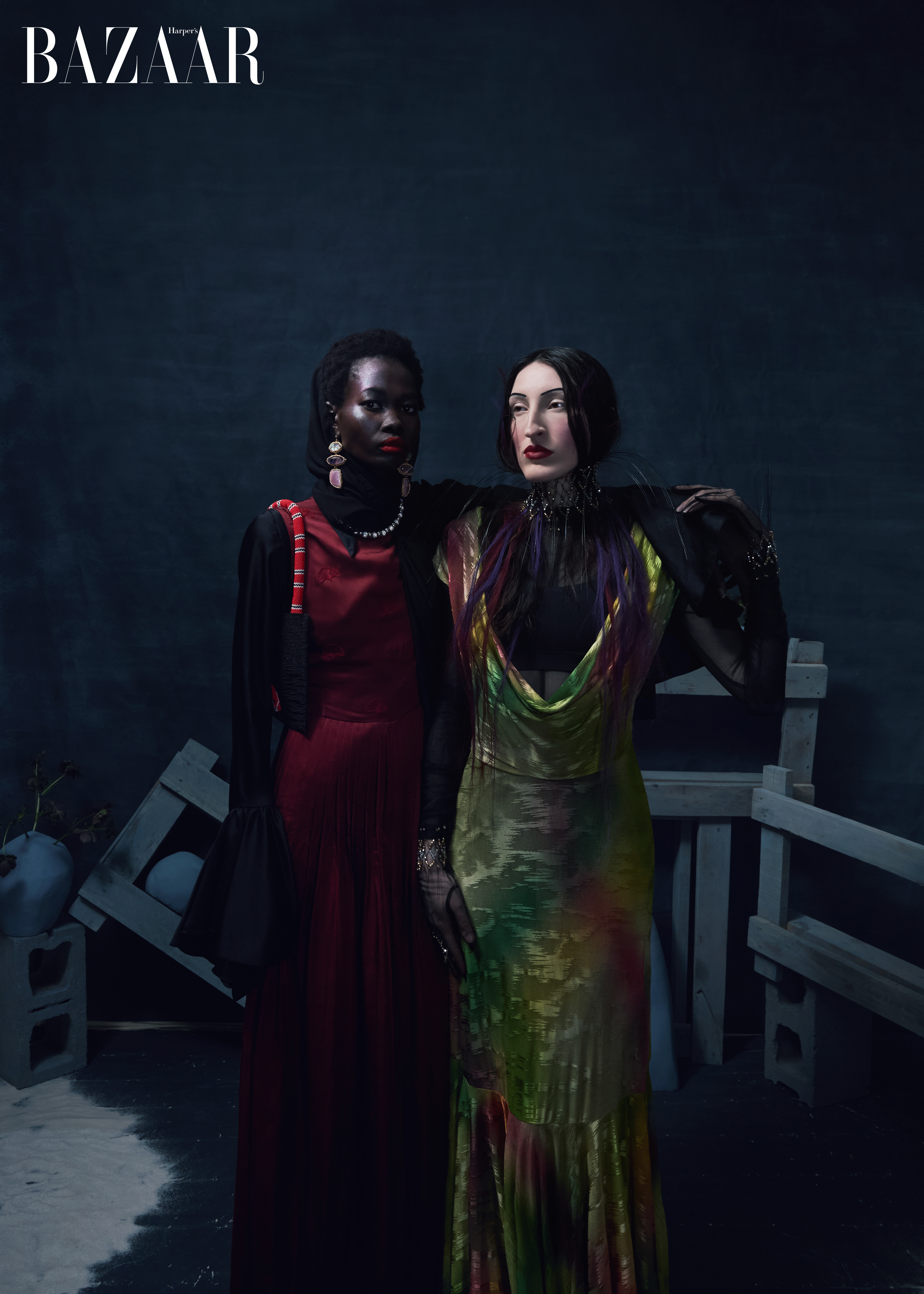 Editorial fashion shot of two adult women in flowing dresses, one red and one patterned, posing indoors in front of a dark background.