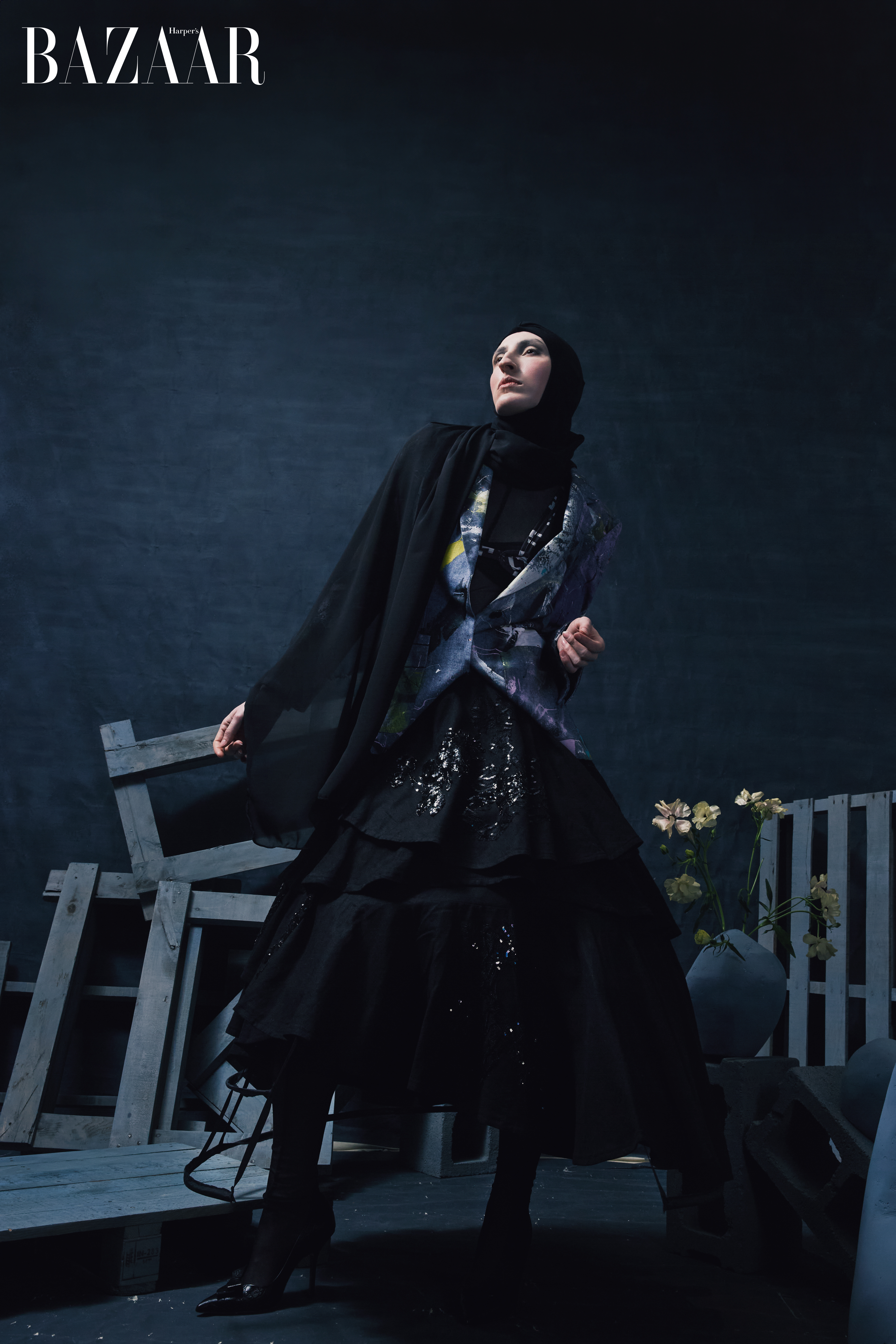 Woman in a black dress and headscarf poses in a dark, moody fashion shot near a wooden pallet and vase of flowers, with BAZAAR logo.