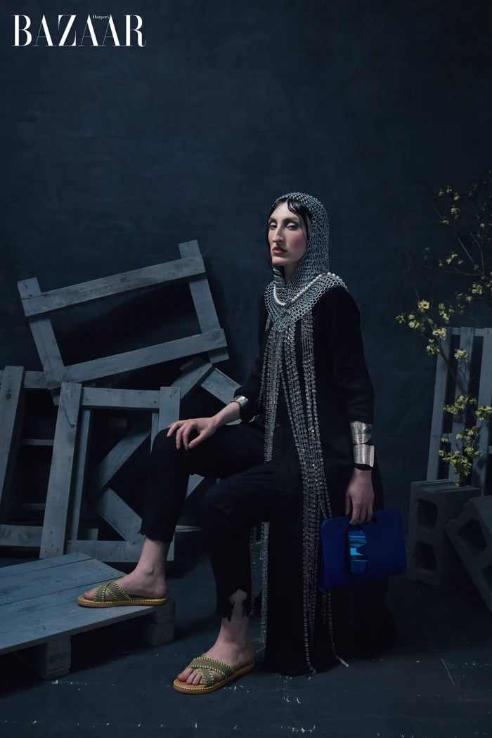 Model wearing a chainmail coif and black coat seated amongst wooden crates for Harper's Bazaar China editorial