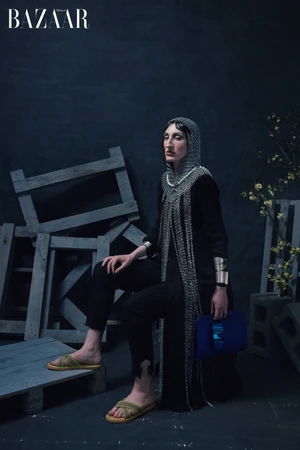 Model wearing a chainmail coif and black coat seated amongst wooden crates for Harper's Bazaar China editorial