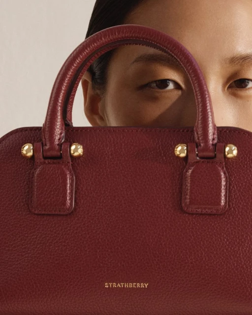 Burgundy Strathberry handbag featuring gold hardware, held in front of an adult with focus on the purse, ideal for fashion accessory search.